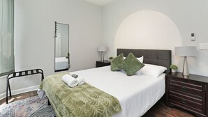 Une chambre d’hôtel bien rangée, avec un grand lit, une table de chevet avec une lampe, un miroir et une petite plante.