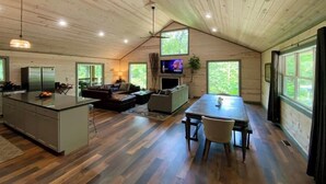 Ein geräumiger Innenraum einer Hütte mit Küche, Essbereich und Wohnzimmer, ausgestattet mit Holzbalkendecken und großen Fenstern.