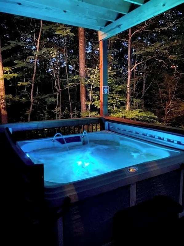 A hot tub under a wooden pergola with a forest backdrop.