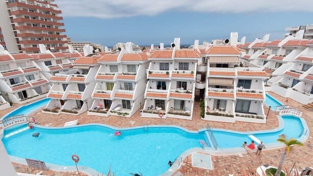 Et hotel med flere hvide bygninger, en central swimmingpool og havudsigt.