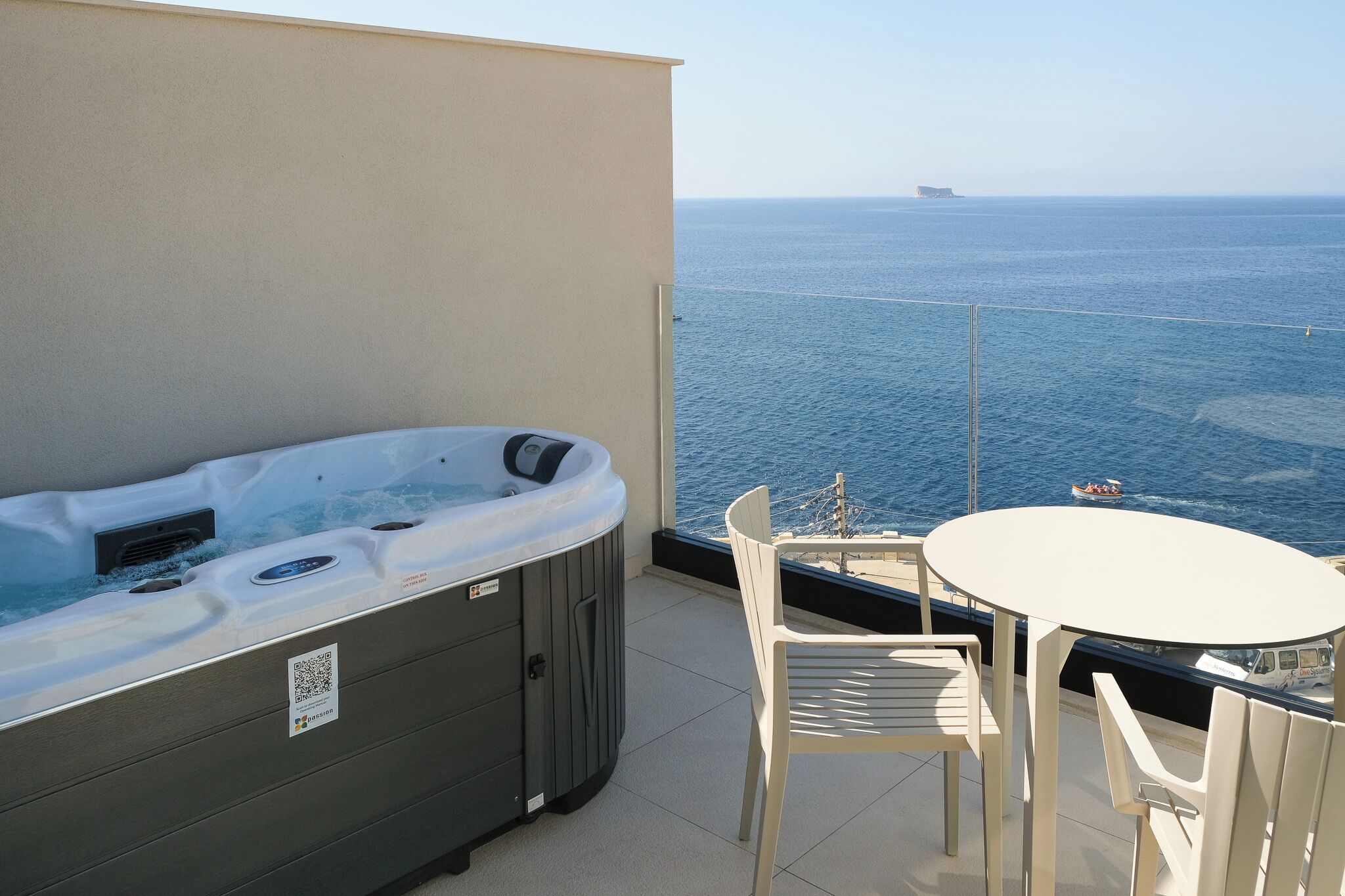 A balcony with a hot tub, a table, and chairs, overlooking the sea.