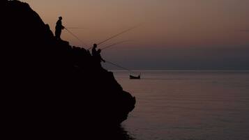 A silhouette of people fishing on a cliff by the sea during sunset.