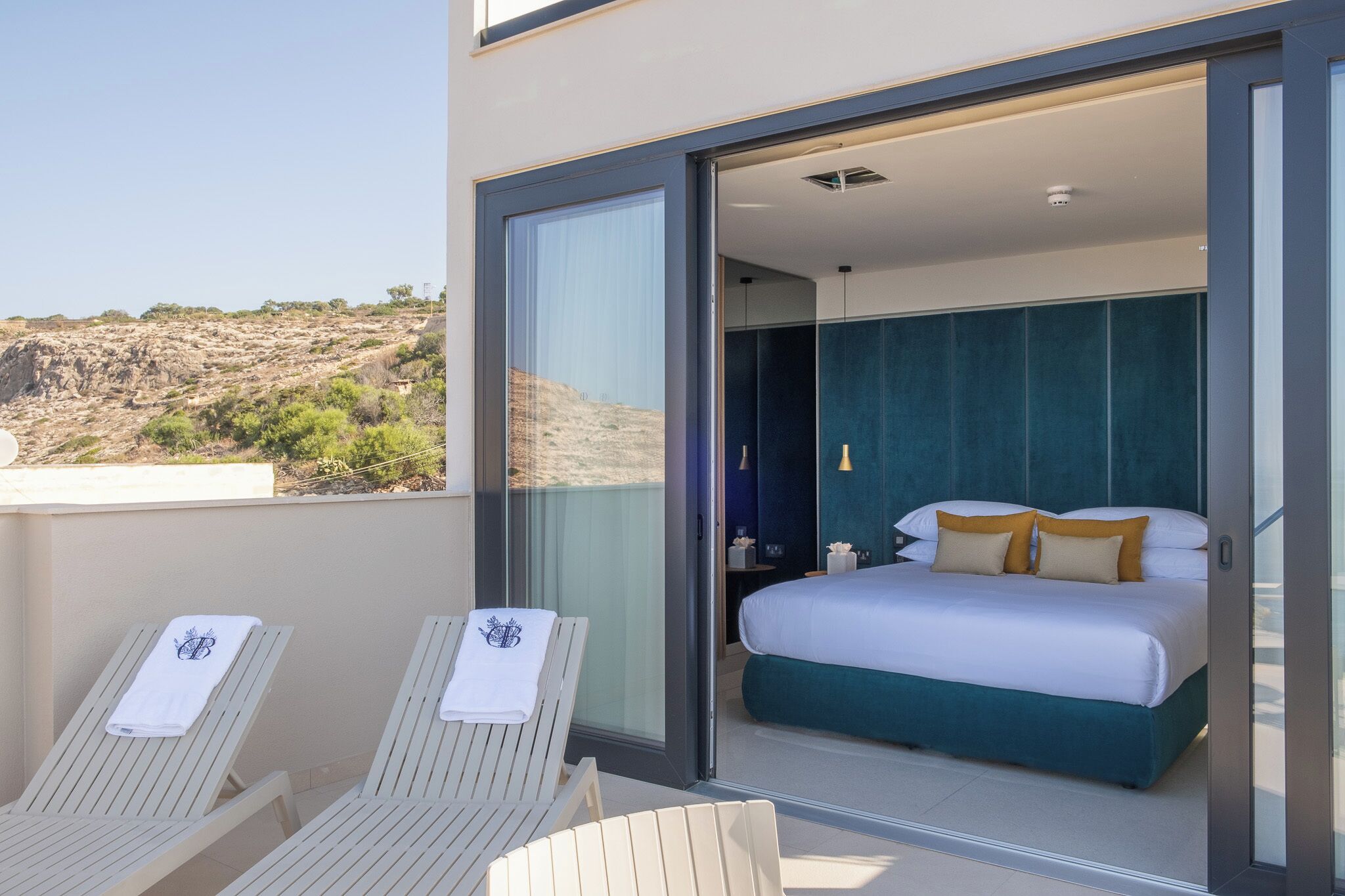Un balcón con dos sillones de descanso y una cama con ropa blanca y cabecera color aguamarina.