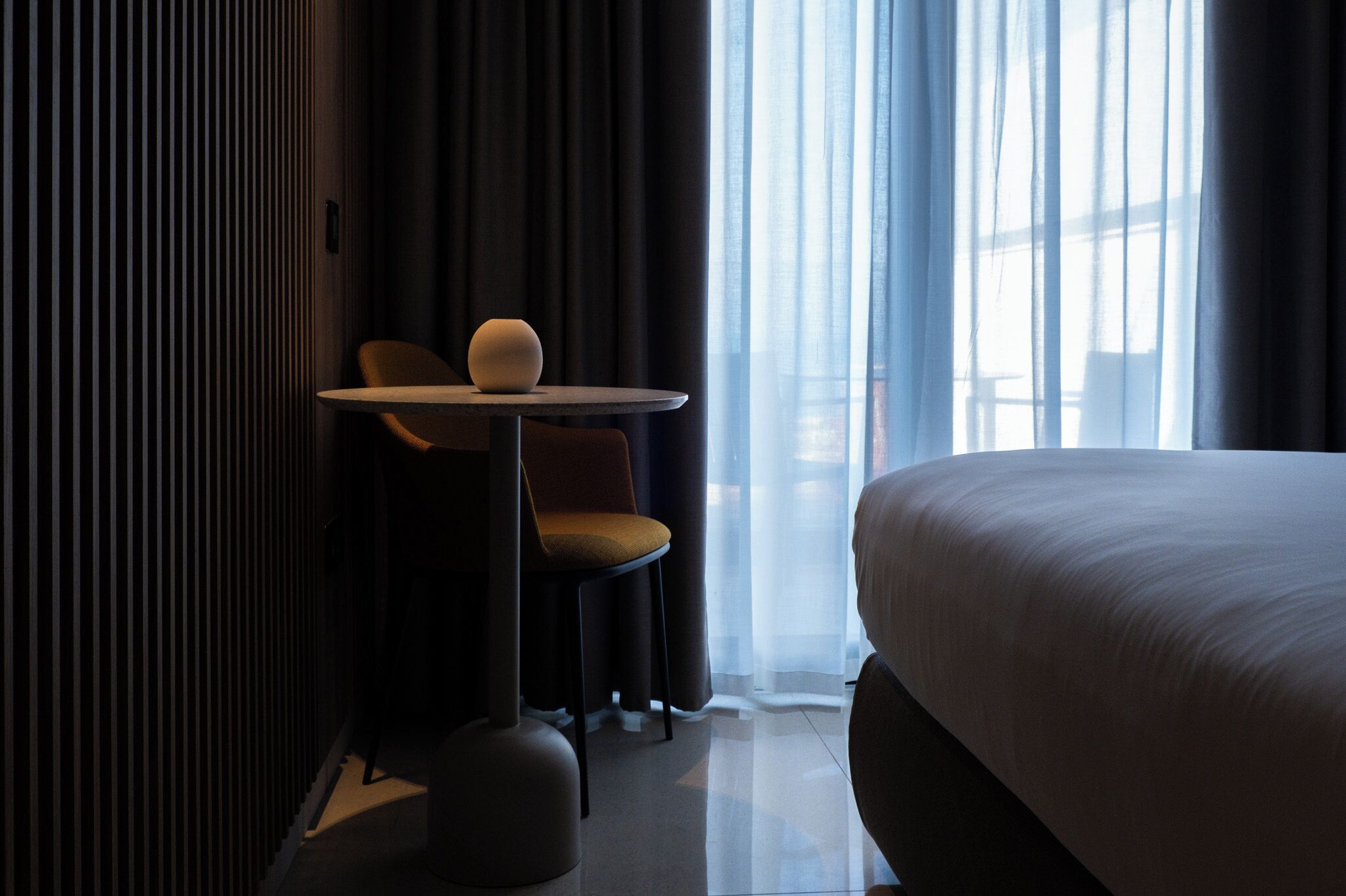 A hotel room with a large bed, a small round table with a chair, and a view of the city through sheer curtains.