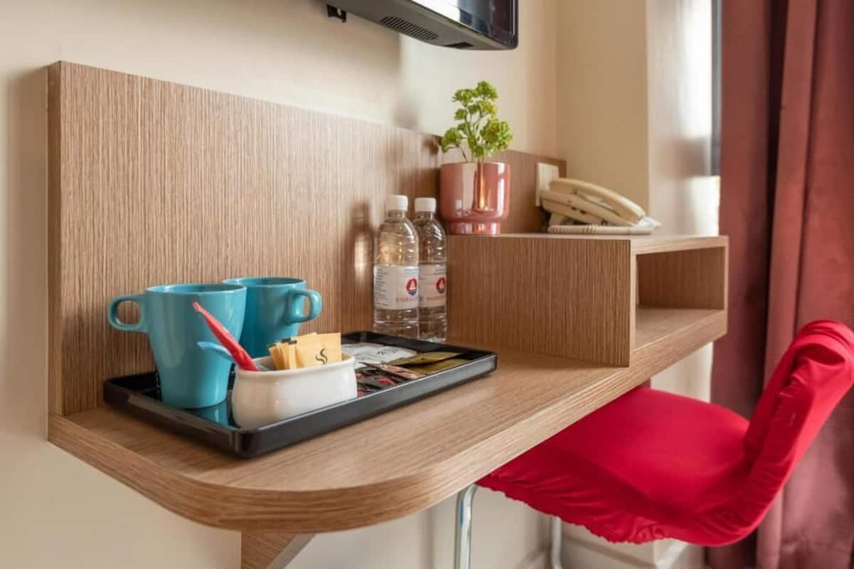 A hotel room with a wooden shelf holding a tray with a blue mug, a white mug, and a tray of snacks. There are two bottles of water and a potted plant on the shelf.