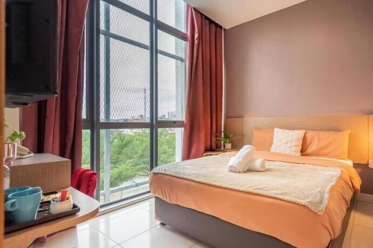 A hotel room with a large bed, a bedside table with a lamp, a TV, and a window with curtains.