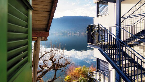 A building with a balcony overlooking a calm lake and distant hills. - Mansarda Verde (Tresa)