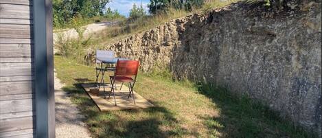 A rustic outdoor seating area with a table and chairs, a stone wall, and a wooden building in the background.
