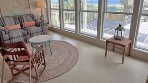A room with a striped sofa, a round table, a rocking chair, and large windows with a view of a balcony and ocean. - Faye DoDo - Beachfront - Island Realty (Grand Isle)