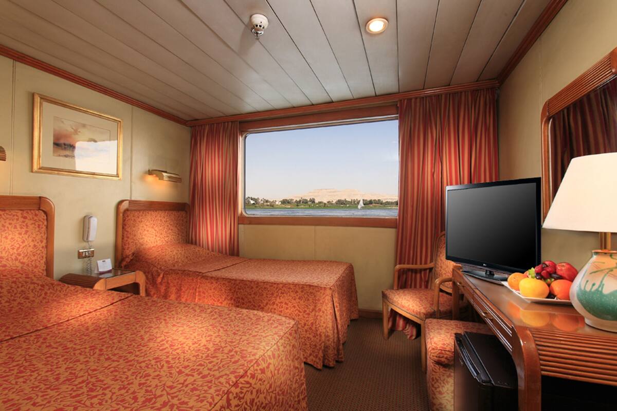 Habitación de hotel con dos camas, televisión, una mesita con fruta y ventana con vista a un paisaje.