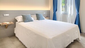 A neatly made bed with white bedding and two blue pillows, a bedside table with a book, and a window with blue curtains.