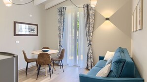 A modern living room with a blue sofa, a dining table with chairs, and a large window with patterned curtains.