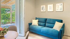 A teal sofa with two pillows, a round table with a bowl of fruit, and a view of greenery through a glass door. - "ULIVO" - Equipped suite, on the ground floor, with veranda. (Levanto)
