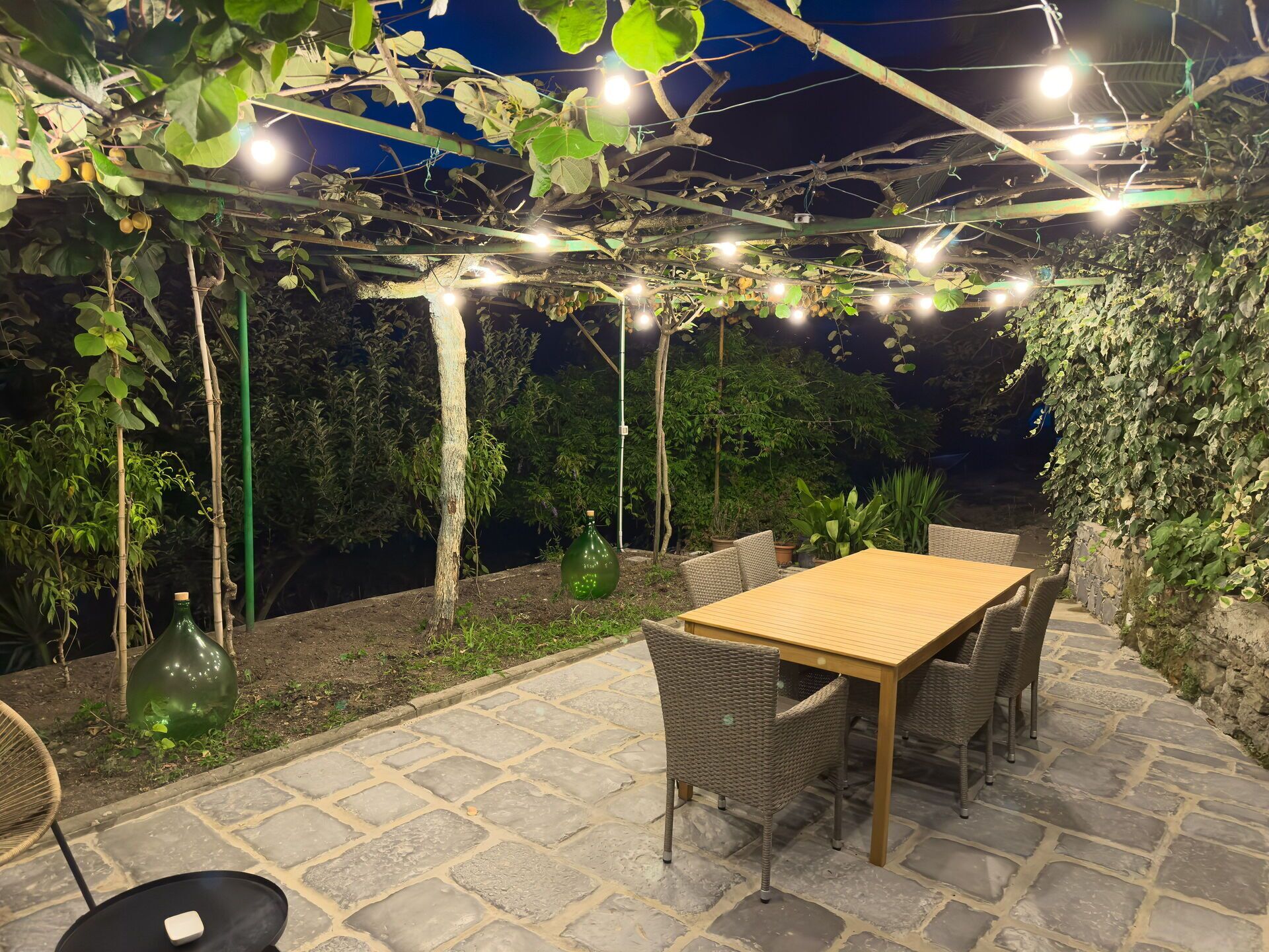 Una terrazza esterna con pavimento in pietra, un tavolo di legno, sedie in vimini e una pergola con luci a stringa.