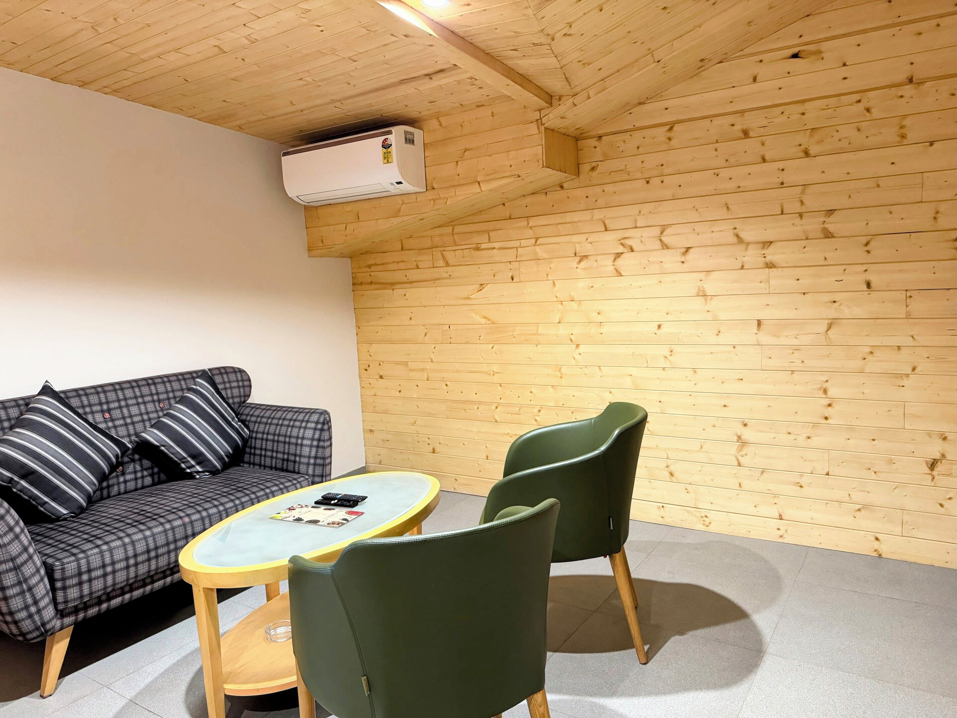 Een kamer met een houten plafond, een bank met gestreepte kussens, een ronde tafel en twee groene stoelen.