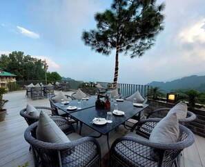 An outdoor dining area with wicker chairs and tables, a flag, and a view of the mountains. - OREN Kasauli  (Kasauli)