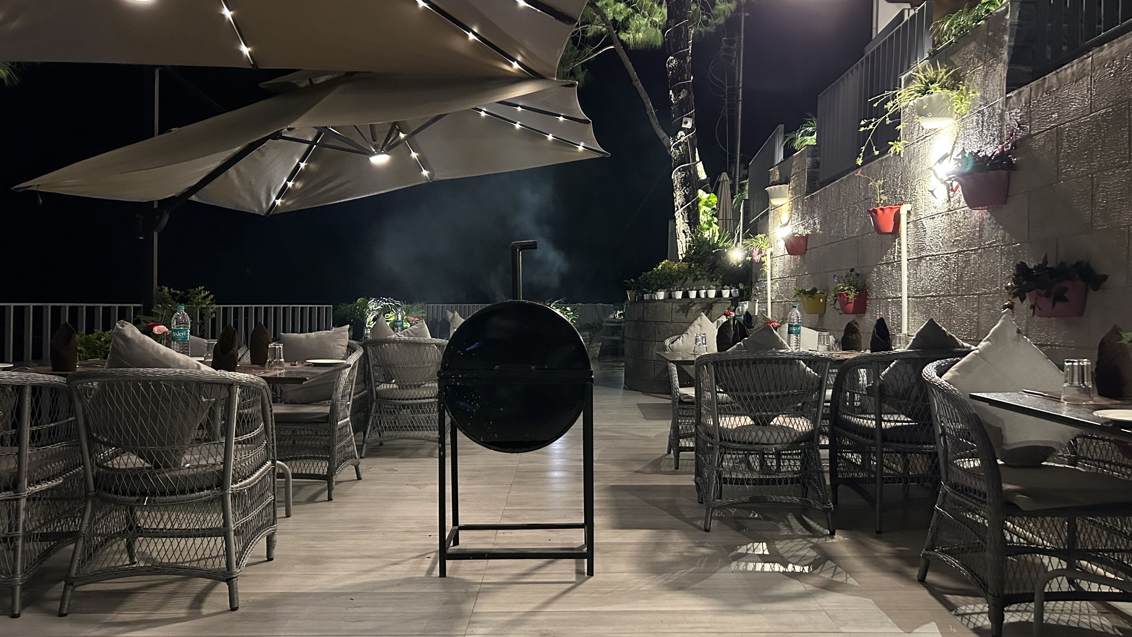 An outdoor dining area with wicker chairs and tables, a large umbrella, and potted plants on the walls.