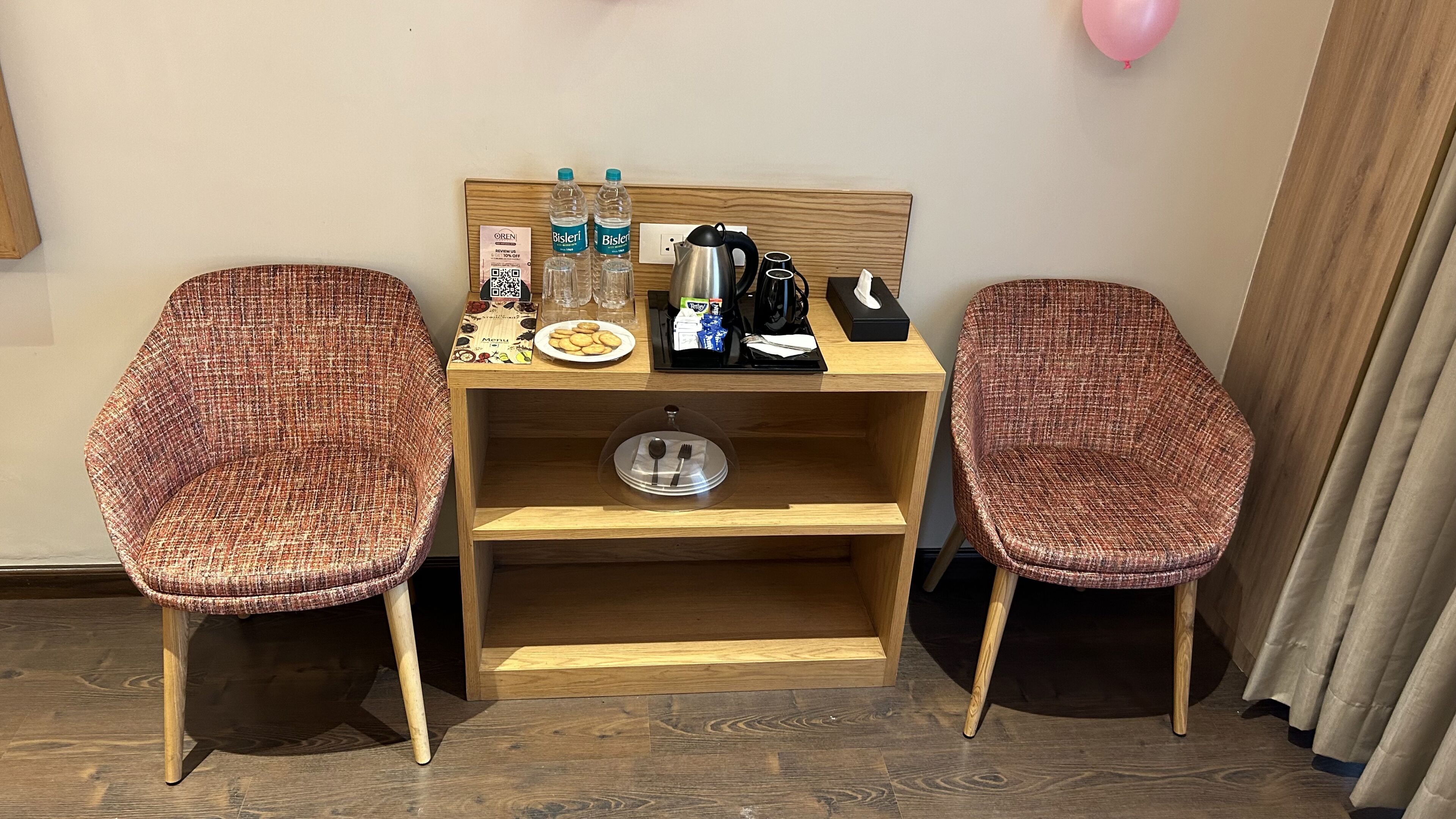 A small, furnished room with two wicker chairs, a wooden desk with a coffee maker, water bottles, and a menu.