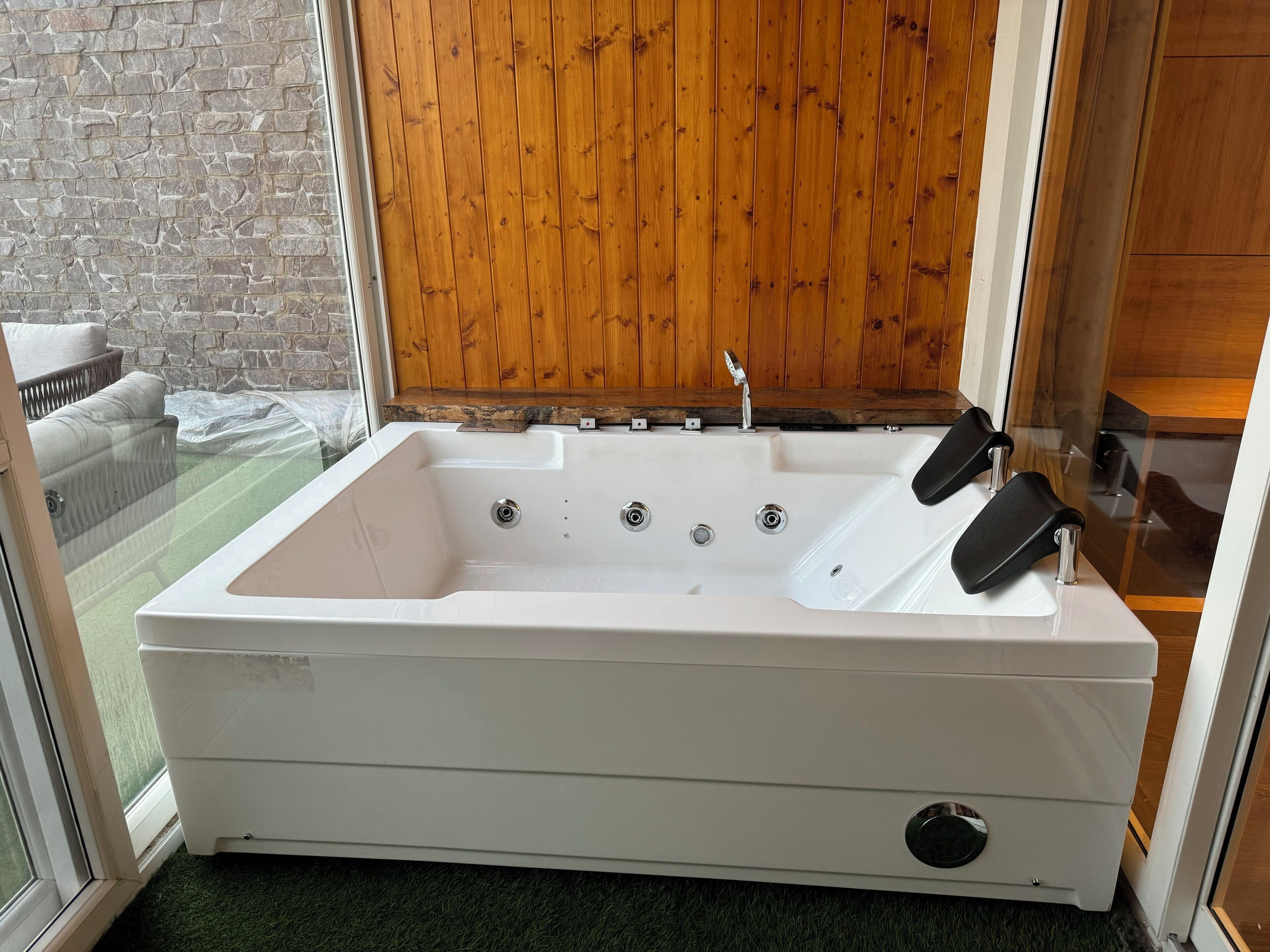 A white spa tub with four faucets, a wooden panel wall, and a glass door leading to a balcony with a white chair.