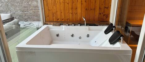 A white spa tub with four faucets, a wooden panel wall, and a glass door leading to a balcony with a white chair.