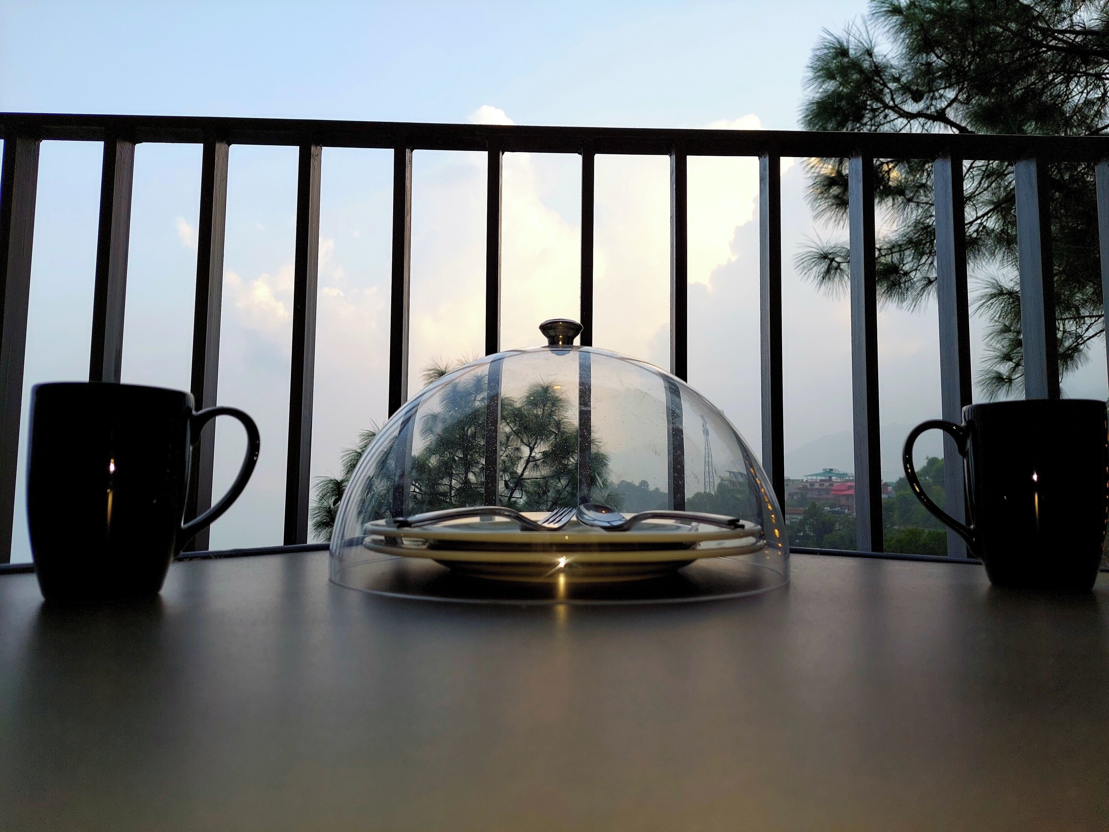 A table with a glass dome cover, two black cups, and a balcony railing.
