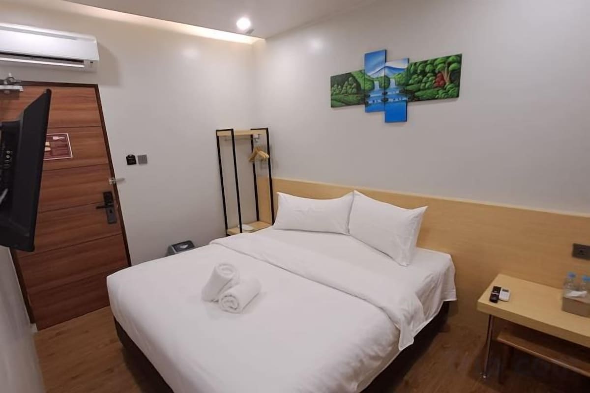 A hotel room with a bed, a bedside table, a TV, and a painting on the wall.