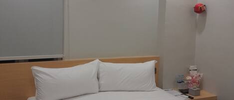 A hotel room with a neatly made bed, a wooden headboard, a bedside table with a red lamp, and a wall-mounted light fixture.