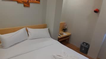 A neatly made bed with white linens and towels arranged in a floral pattern, a wooden headboard, a small bedside table with a lamp, and a painting of a sunset on the wall.