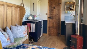 A cozy room with a bed, a small kitchenette, a wooden door, and a red stove. - Shepherd Hut in Private Meadow With Outdoor Bath (Langport)