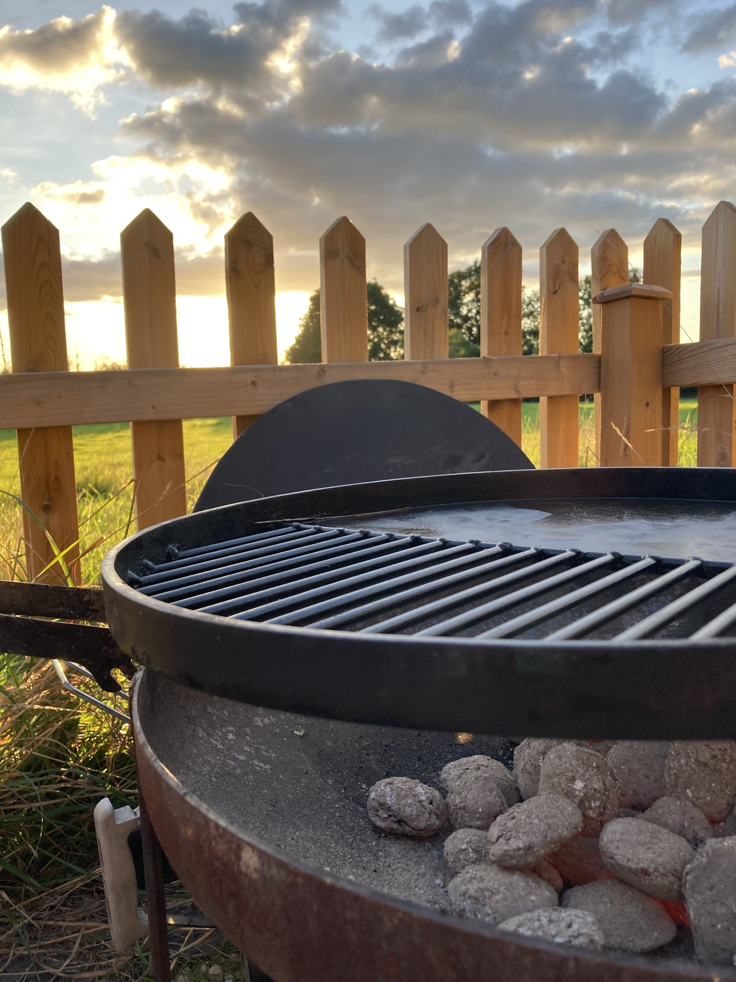 A barbecue grill with charcoal, a wooden fence, and a cloudy sky in the background.