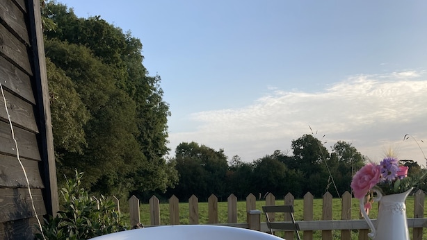 A white hot tub filled with water and rose petals, surrounded by a wooden fence and greenery. - Shepherd Hut in Private Meadow With Outdoor Bath (Langport)