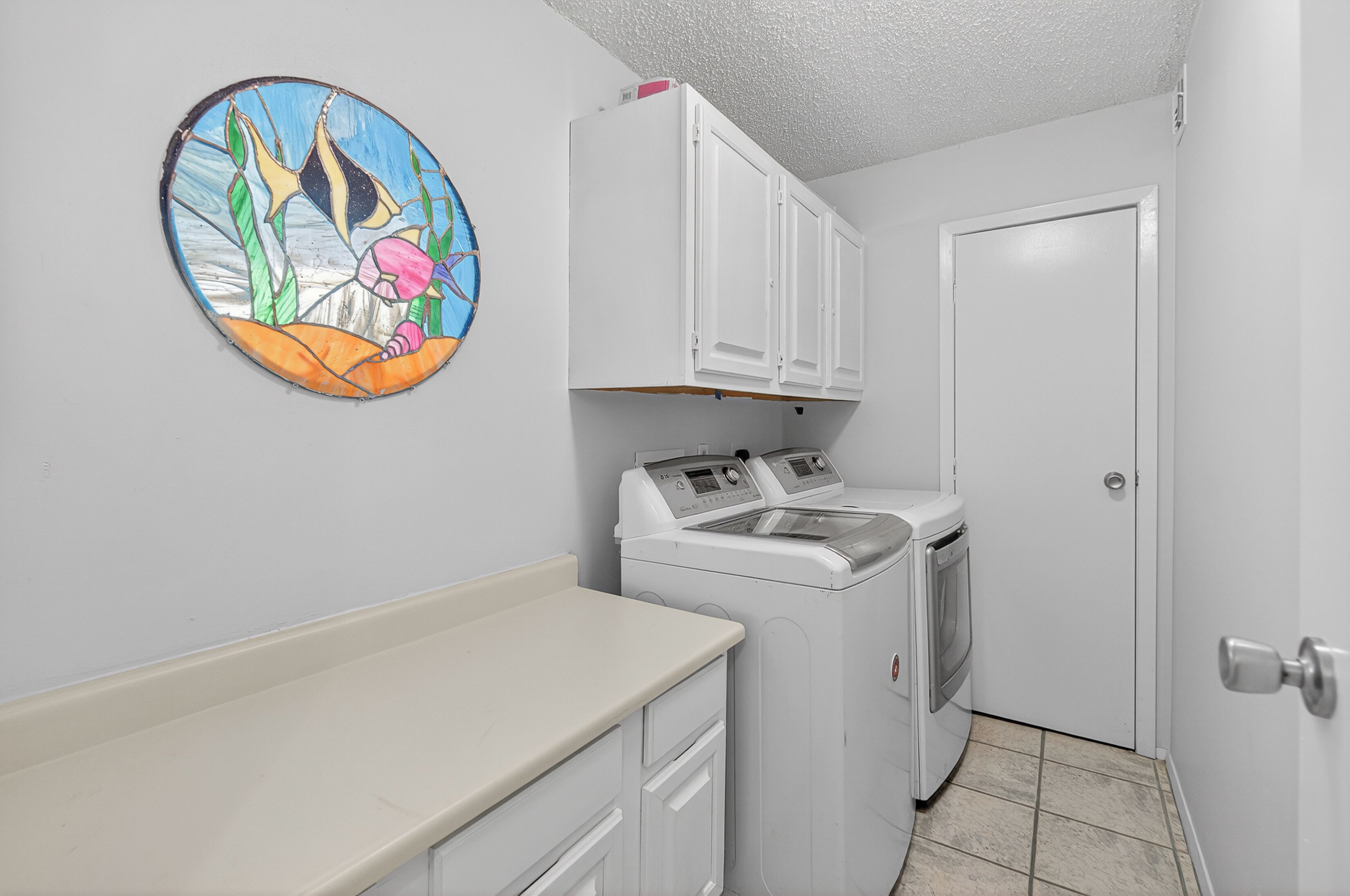 A laundry room with a washing machine, a dryer, and a cabinet above the machine. There is a door to the right and a wall-mounted artwork on the left.