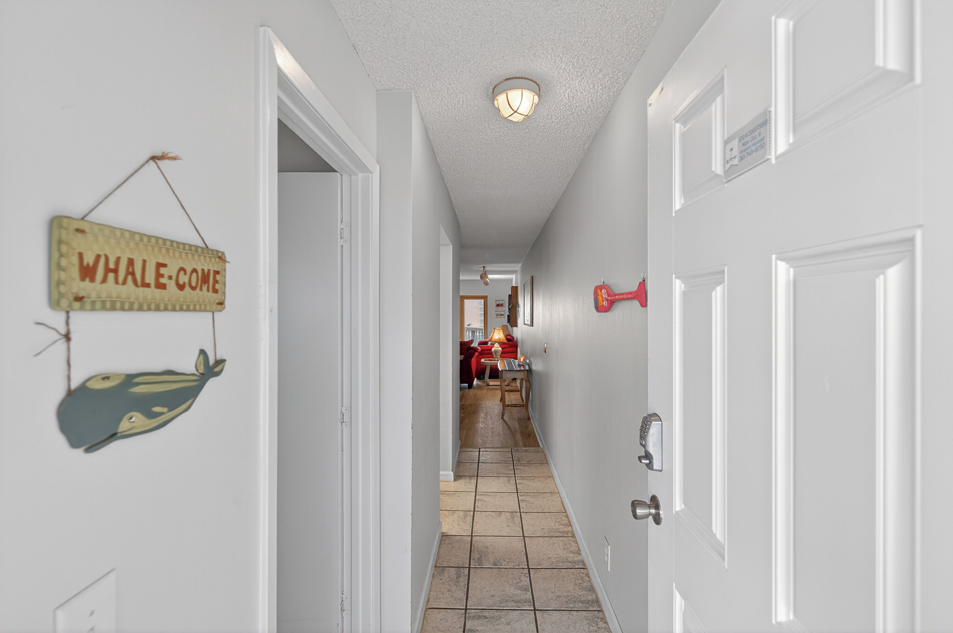 A hallway with white walls, a tiled floor, and a door with a red handle. A sign on the wall reads "WHALE-COME."