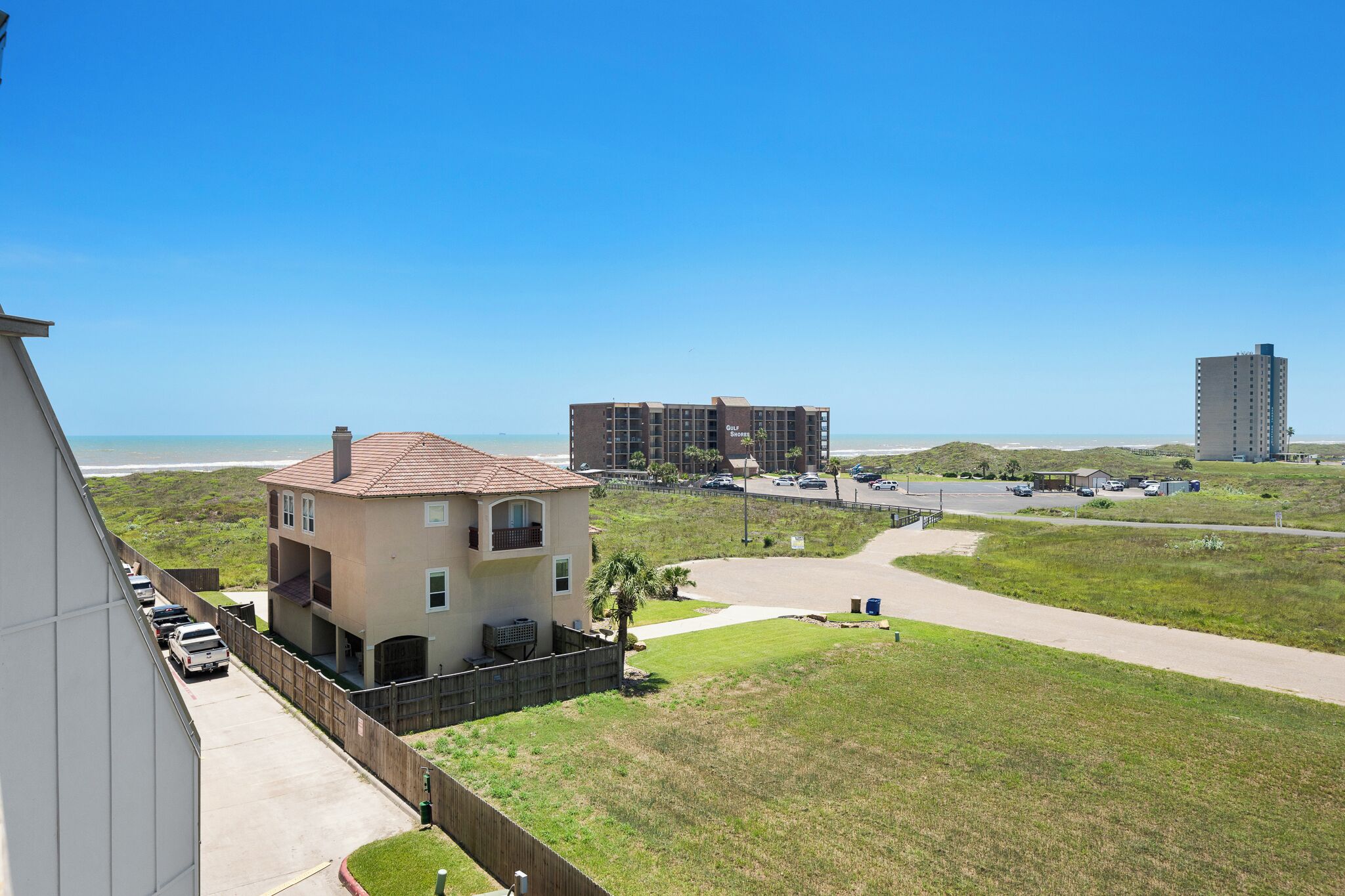 A coastal residential area with a two-story house, a parking lot, and a beach in the background.