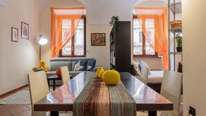 A dining area with a long table, chairs, and a sofa. There are two windows with orange curtains, a bookshelf, and a lamp. - Carignano 8 - Turin Central Loft Carignano (Turin)