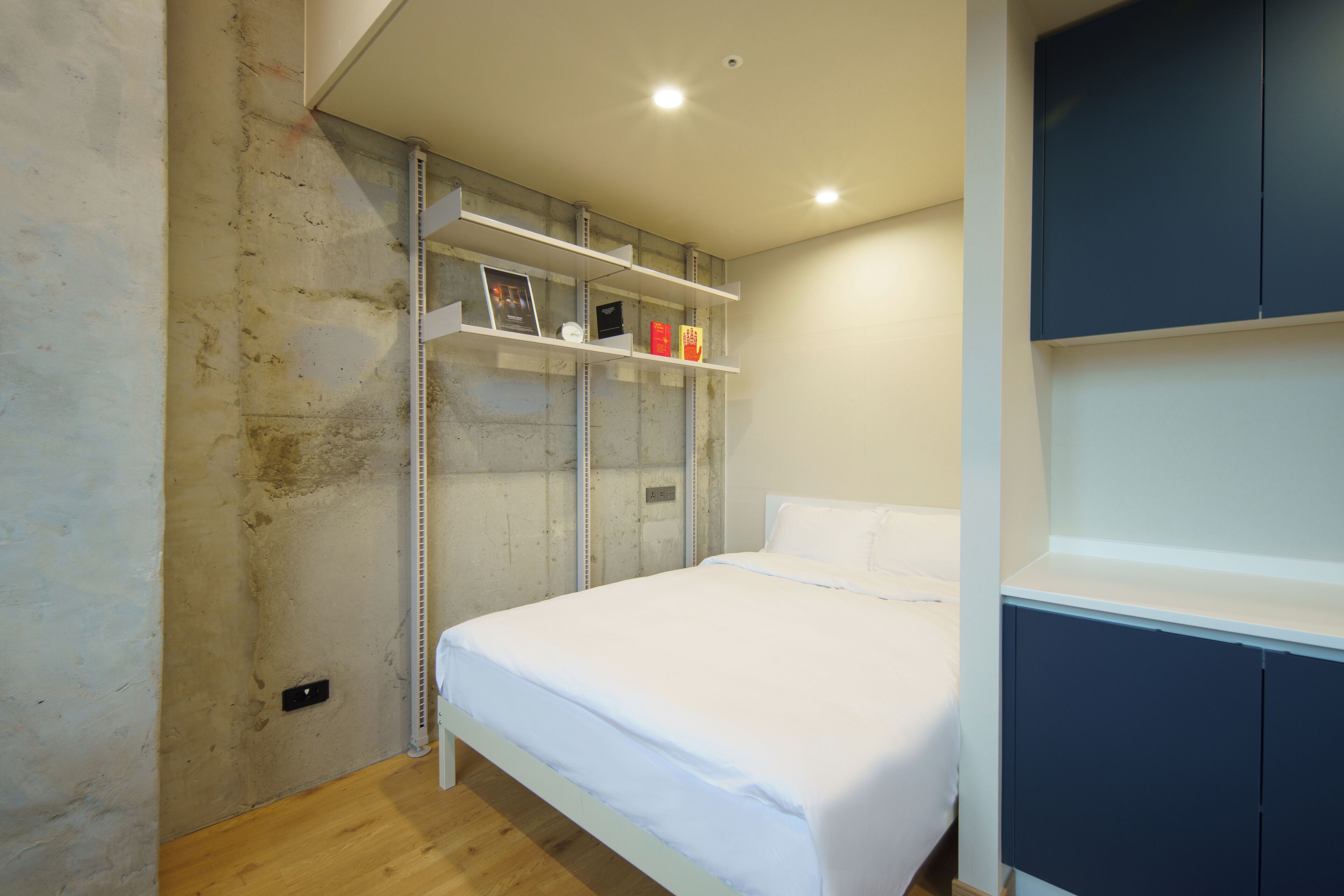 Een slaapkamer met een bed, witte beddengoed, een plank met boeken en een garderobe.