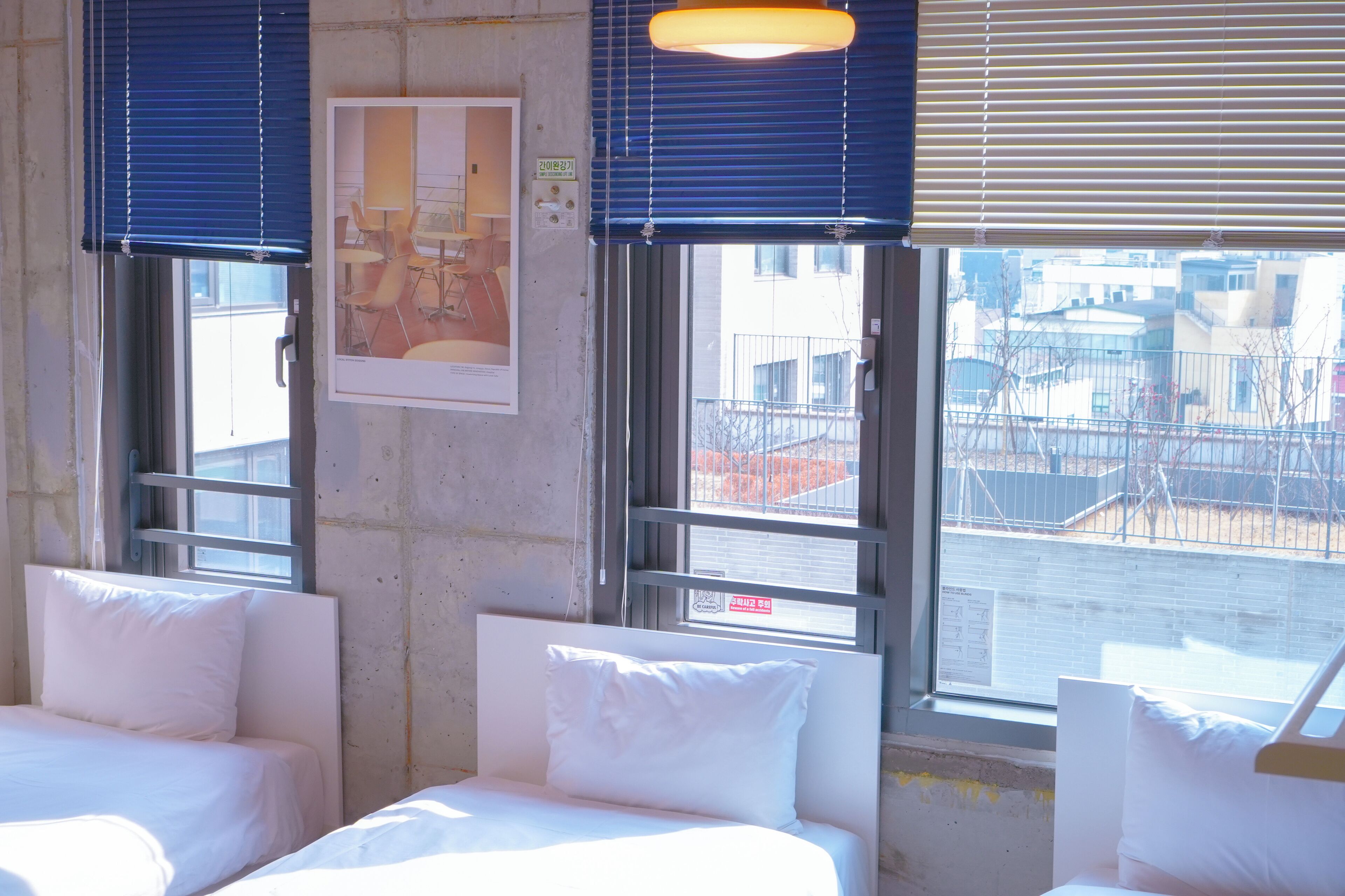 Habitación de hotel con dos camas, ventanas amplias con persianas y una vista del paisaje urbano. En la pared, un cuadro enmarco.