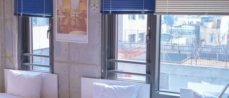 Habitación de hotel con dos camas, ventanas amplias con persianas y una vista del paisaje urbano. En la pared, un cuadro enmarco.
