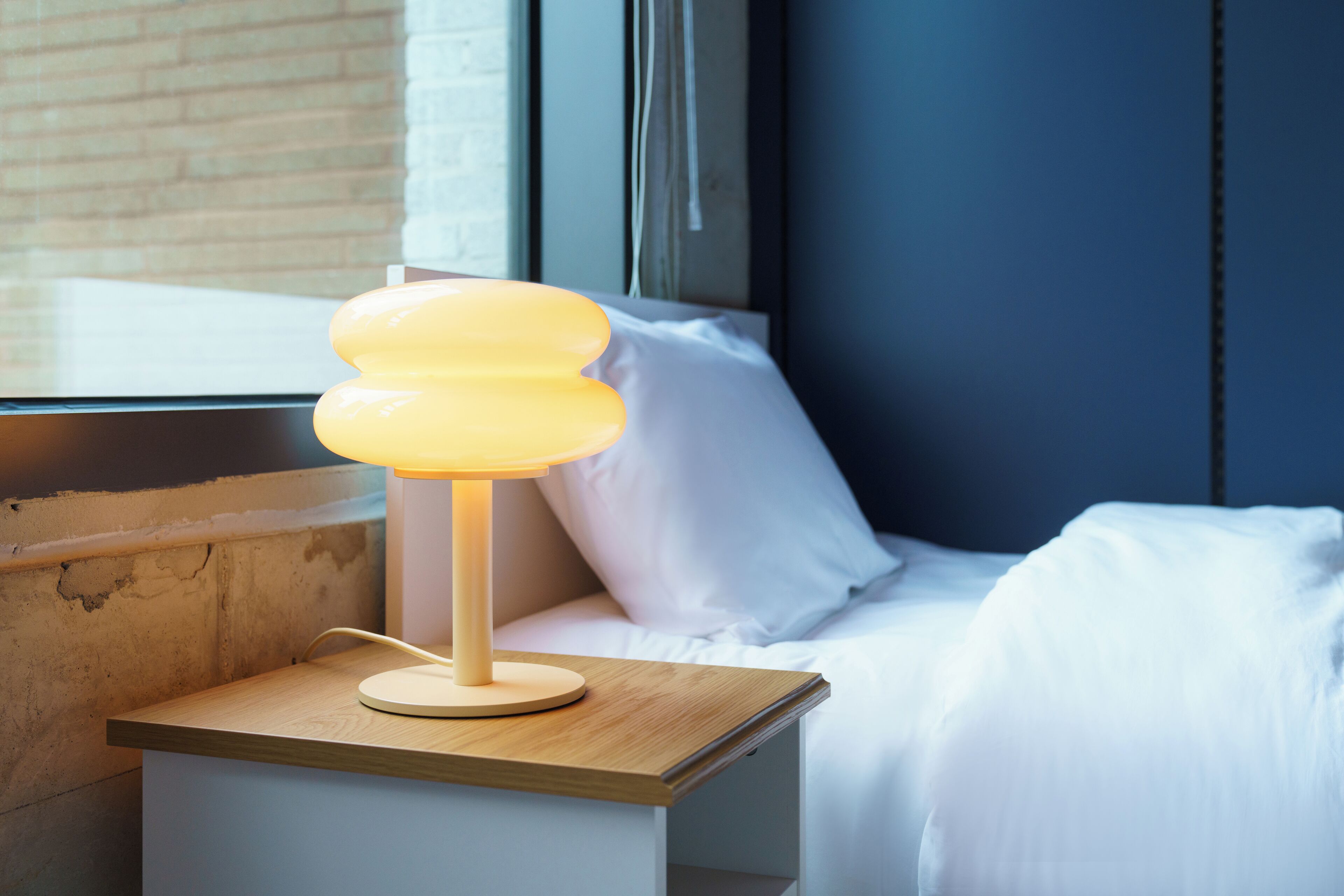 Une table de chevet avec une lampe, un lit avec du linge de lit blanc et une fenêtre avec des stores.