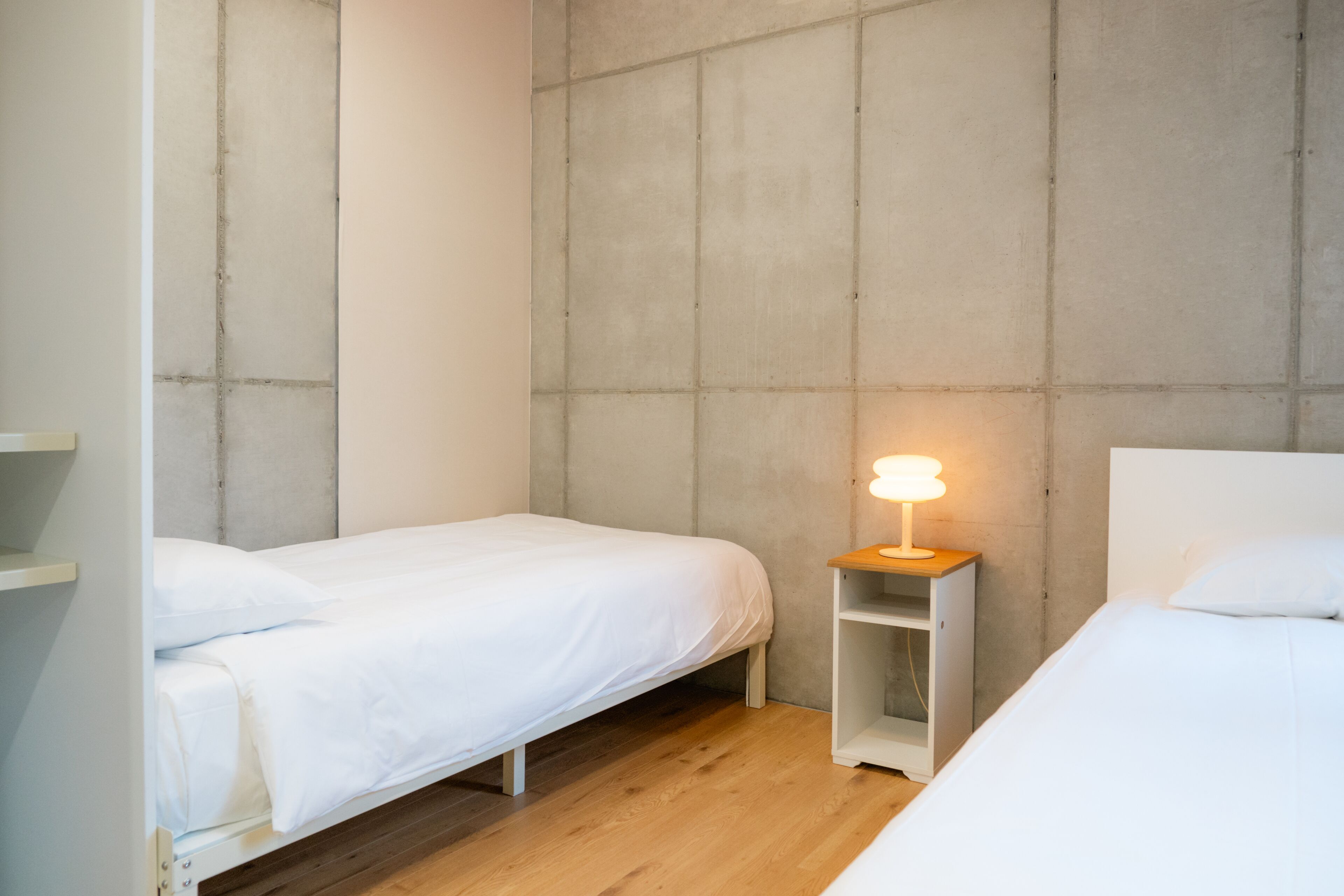 Una habitación pequeña y moderna con dos camas individuales, una mesita de noche con lámpara y una pared de concreto decorativa.