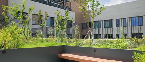 An outdoor seating area with a bench, surrounded by greenery and a modern building with large windows.