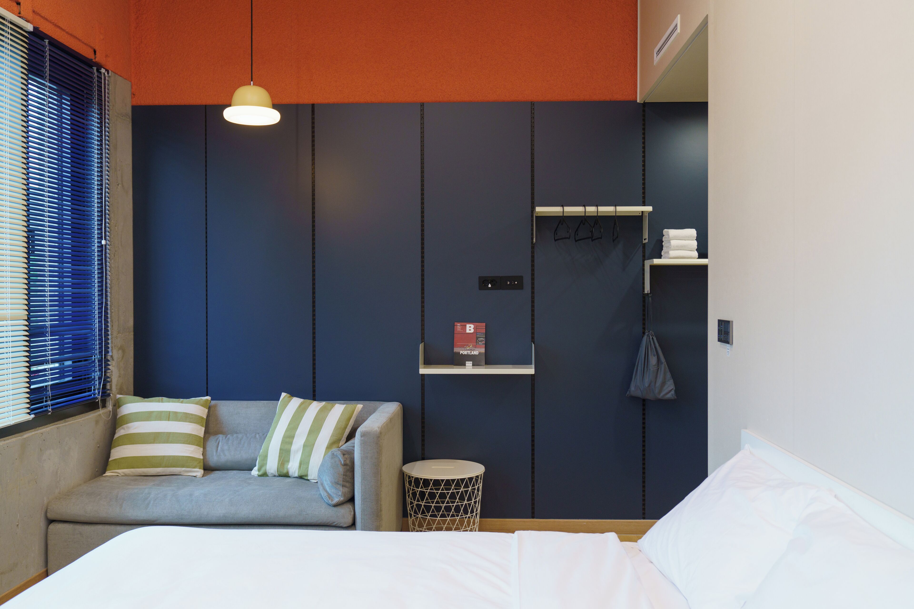 A modern hotel room with a grey sofa, a small round side table, a bed with white linens, a hanging bag, and a shelf with a book and a towel.