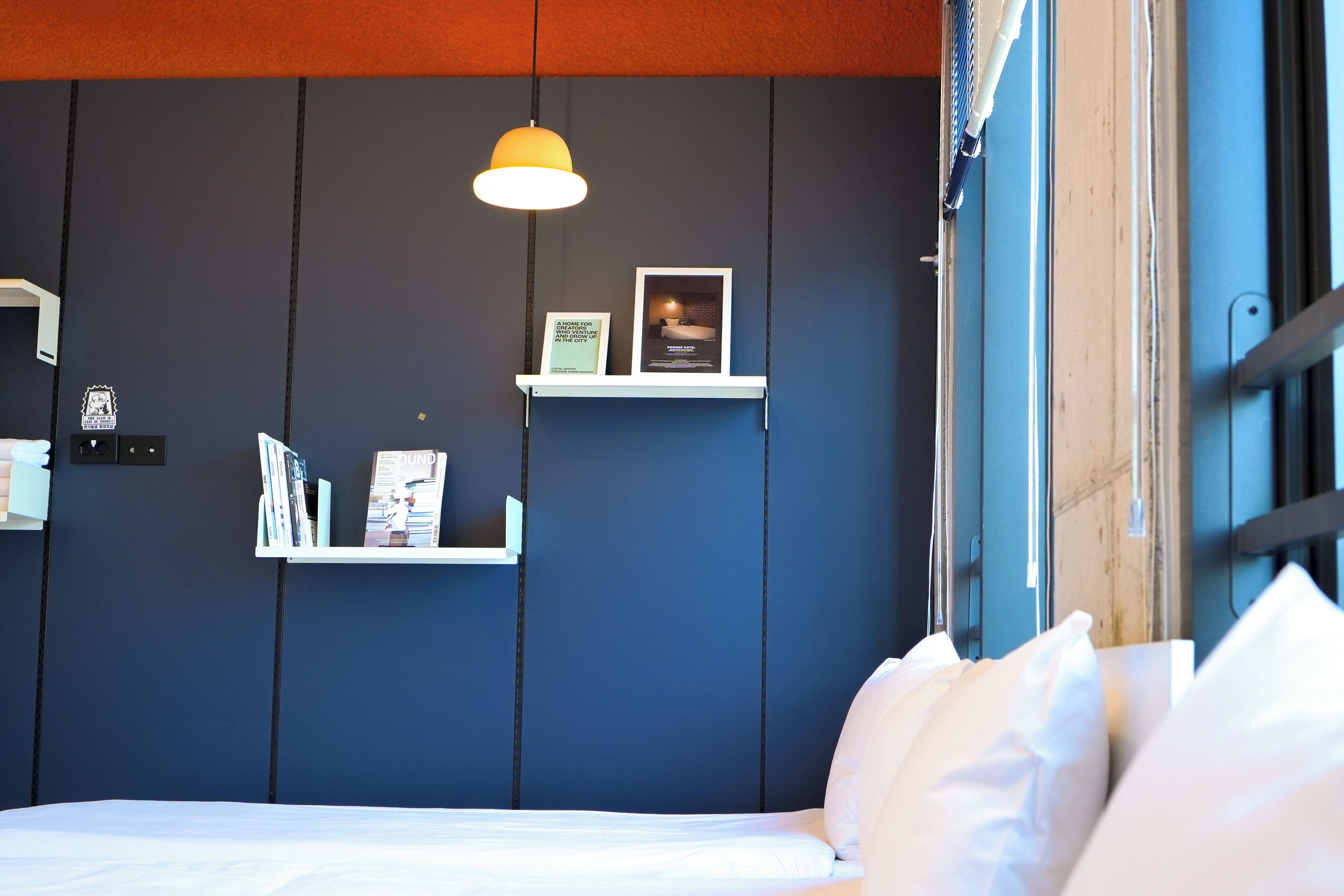 Une chambre à coucher avec un lit, des oreillers blancs, un luminaire suspendu et deux étagères blanches ornées de livres et de photos encadrées.