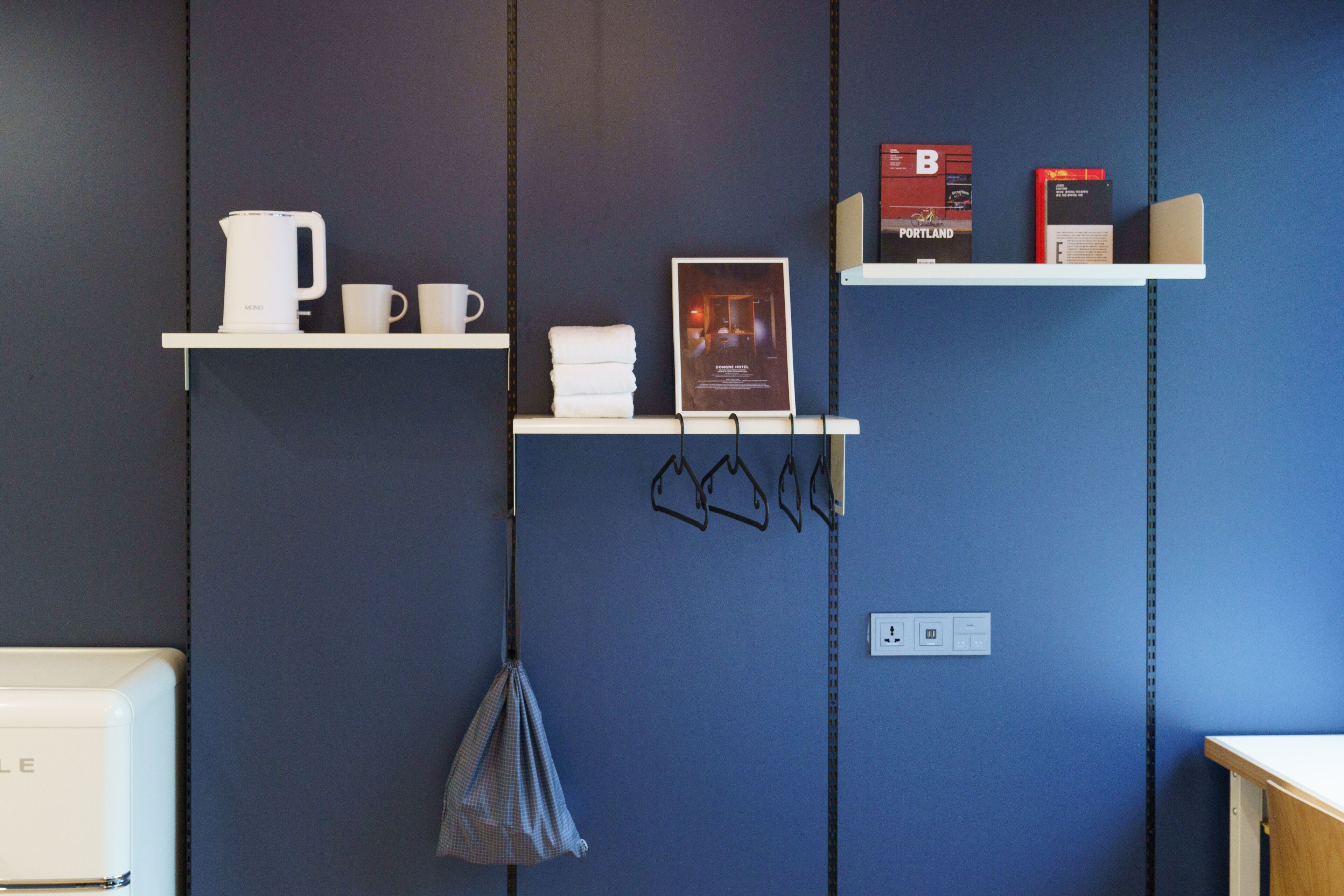 Habitación con dos estantes blancos contra una pared azul, cada uno con objetos como una tetera, tazas, un cartel y libros.