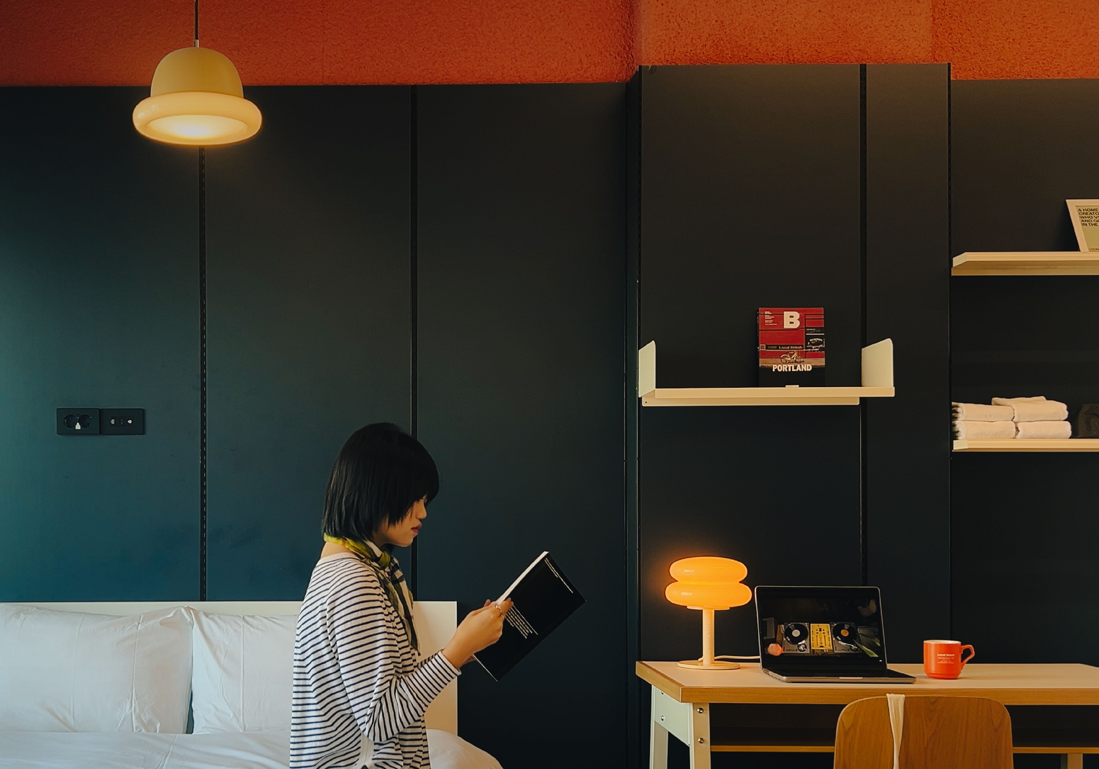 A room with a bed, a desk with a laptop, a lamp, and a shelf with books and a red box.