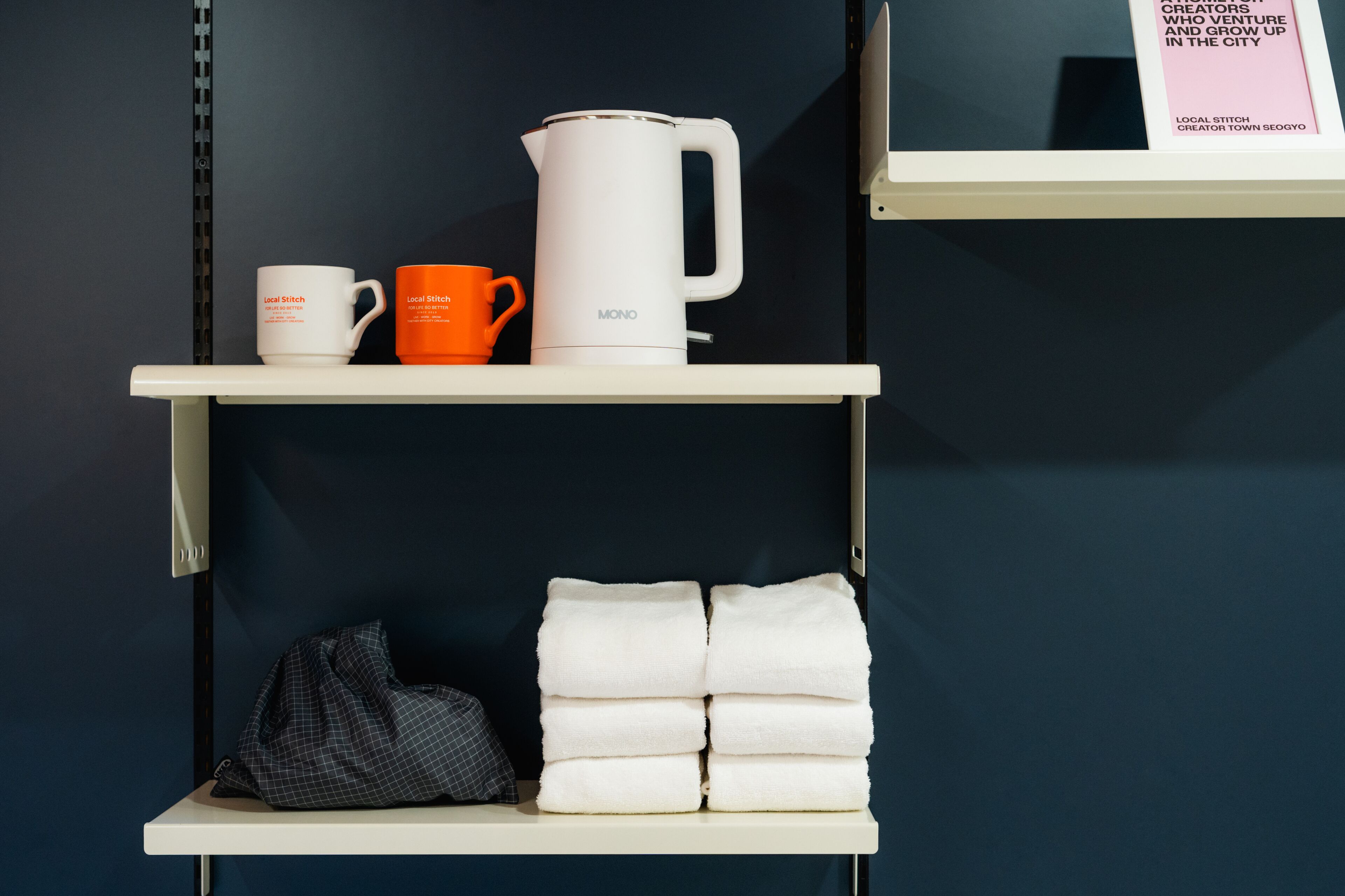 A shelf with a white mug, an orange mug, and a white kettle. Below, there are folded white towels and a black bag.