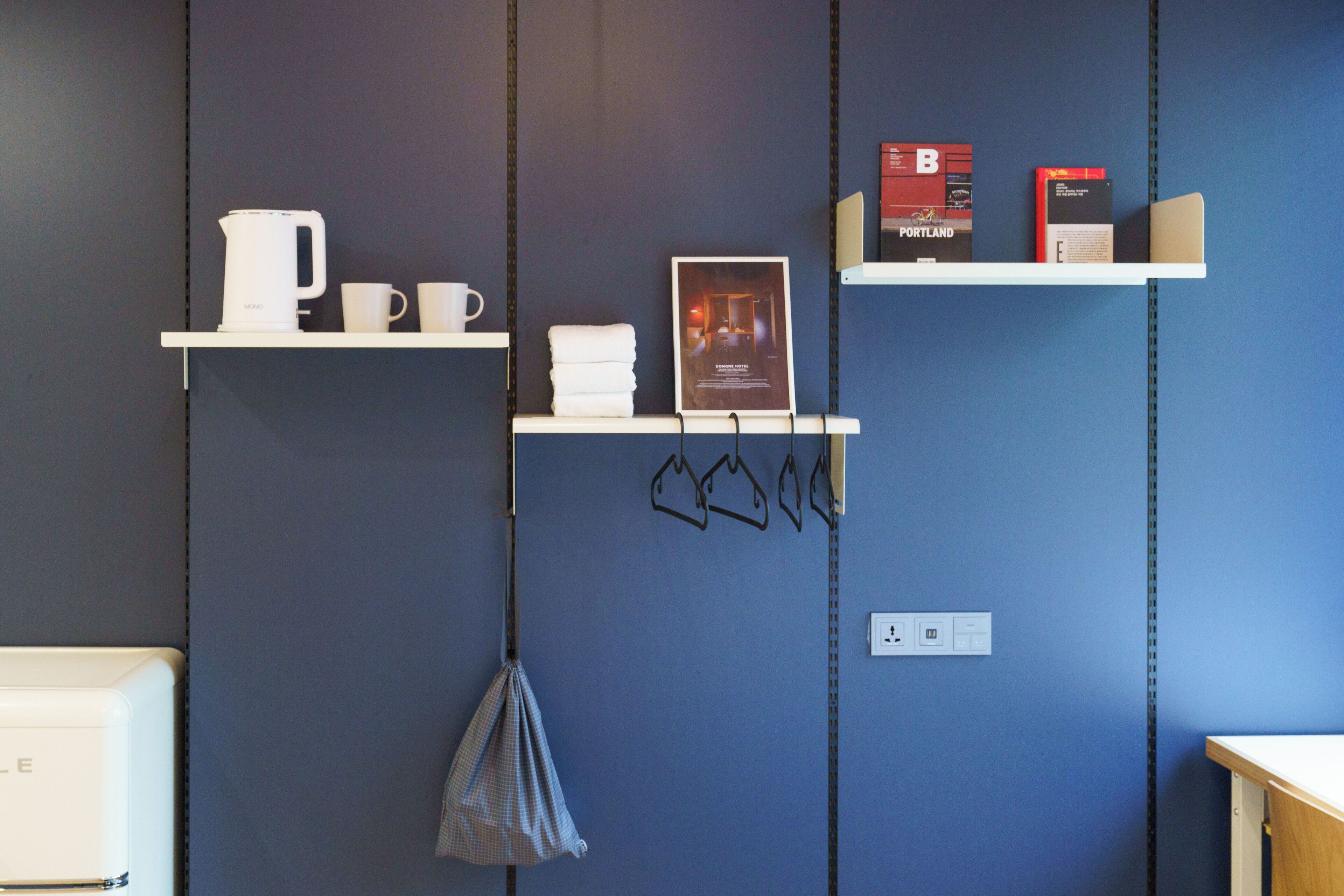 Une pièce avec un mur bleu, deux étagères blanches sur lesquelles sont disposés divers objets, une bouilloire blanche, des tasses, une affiche et un sac suspendu.