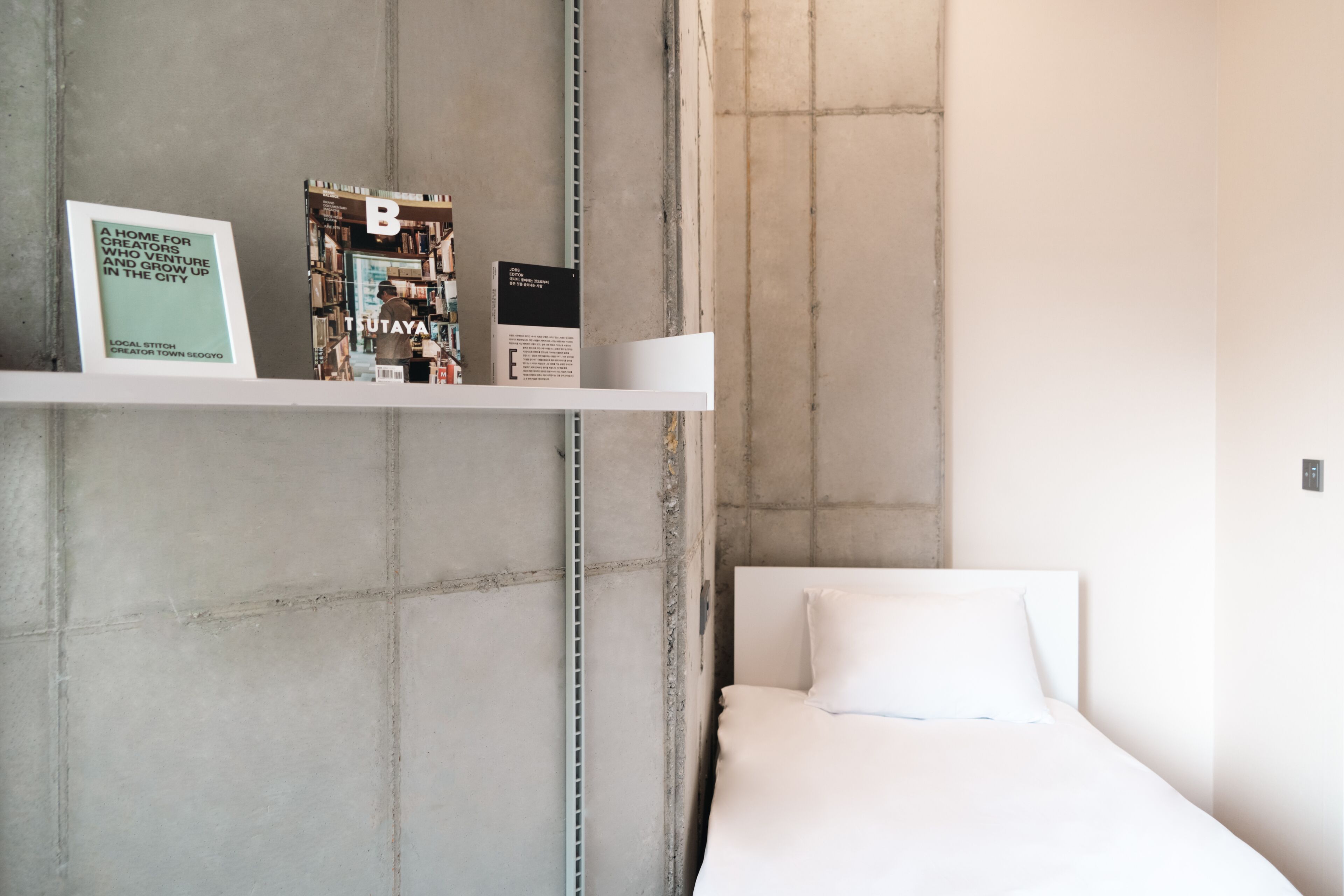 Um quarto com uma cama, uma estante branca com livros e uma placa, e uma parede de concreto.
