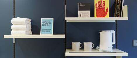 Two white shelves against a blue wall, one holding folded white towels and a sign, the other with two white mugs and a white electric kettle.