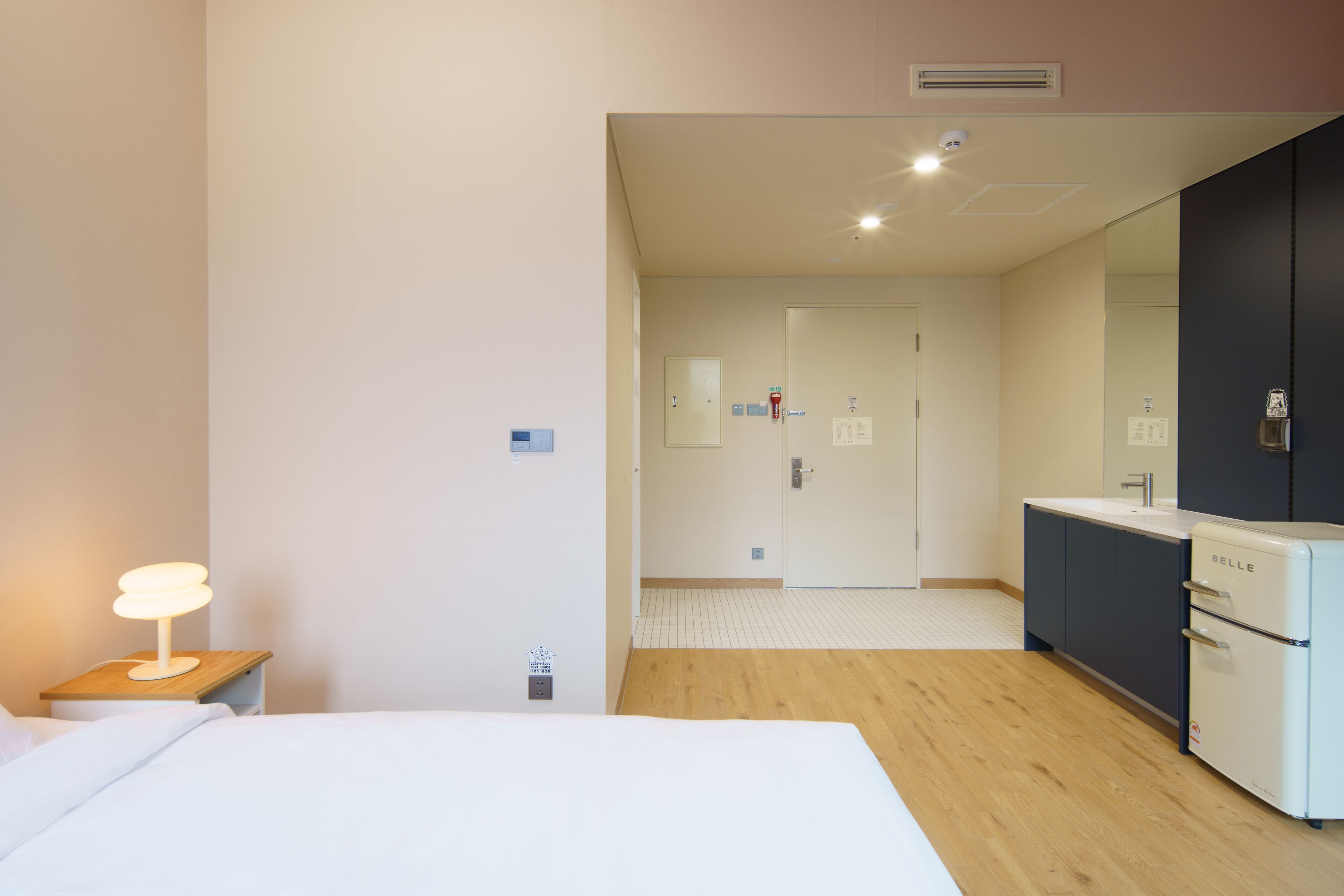 A hotel room with a bed, bedside table with a lamp, a large mirror, a kitchenette with a fridge, and a door leading to another room.